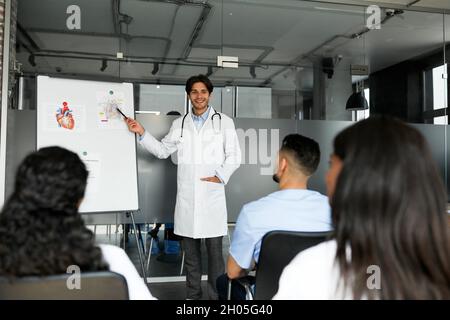 Cheerful young man chief doctor standing by presentation board, having speech, multiracial team of professional doctors attending medical conference o Stock Photo