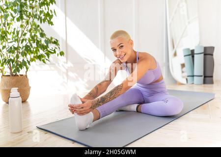 Domestic sports concept. Positive young woman in sportswear doing leg stretch exercises, sitting on mat on floor in living room and smiling to camera, Stock Photo