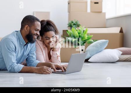 Smiling young african american woman and man looking at laptop with new design for room, lying on floor with boxes and plants, working on interior sty Stock Photo