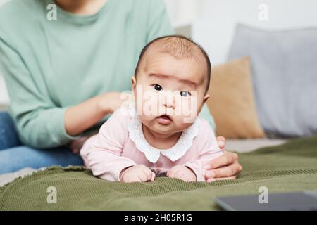 Portrait of Asian baby girl looking at camera while lying on the stomach on the bed with mother holding her in the background Stock Photo