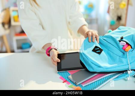 Closeup on school girl with tablet PC, workbooks and blue backpack packing for school at home in sunny day. Stock Photo