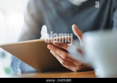 Hand of a Black man holding a tablet pc in close up viewed past a mug of coffee on a table Stock Photo