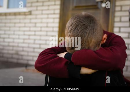 Sad little boy sitting on stairs near his house with closed door. Child cry and hiding his face. Kid alone. Victim of bulling. Problem in school. Soci Stock Photo