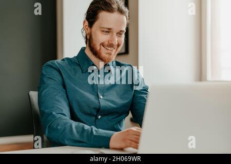 Smiling young male office worker typing on keyboard and looking at laptop screen in office. Stock Photo
