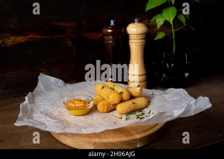 A portion of cheese sticks, served on a wooden board with mustard. Bar deep fried food. Shot in the interior of a bar against a dark wooden background Stock Photo