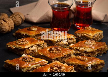 Honey baklava with walnuts on black slate. Portions of oriental sweets. Turkish sugary pieces. Tea poured in armudu glass in the background. Stock Photo