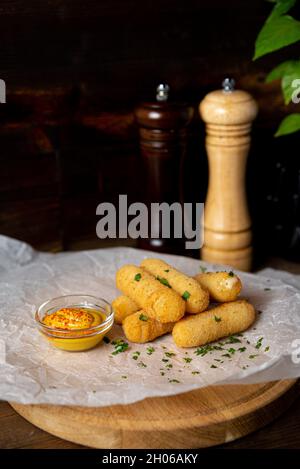 A portion of cheese sticks, served on a wooden board with mustard. Bar deep fried food. Shot in the interior of a bar against a dark wooden background Stock Photo