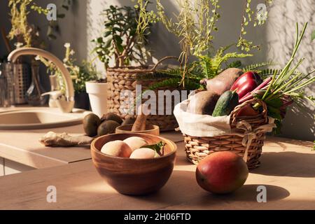 Basket with fresh vegetables surrounded by fruits and domestic plants in the kitchen Stock Photo