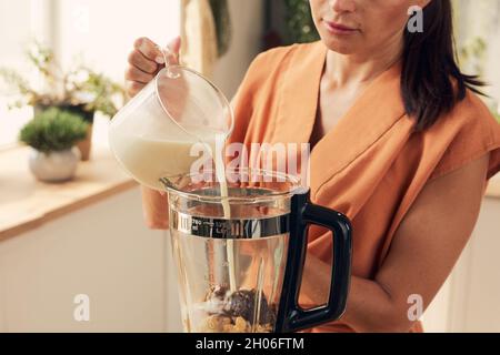 Young female in casualwear pouring fresh milk into electric blender while making smoothie Stock Photo