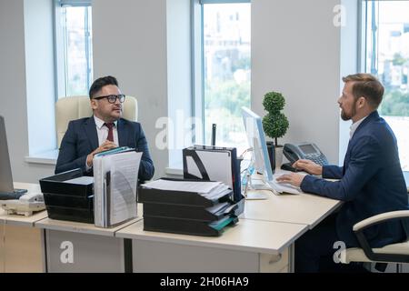 Two contemporary businessmen interacting while sitting by their workplaces Stock Photo