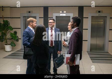 Group of young intercultural colleagues in formalwear interacting in corridor of large business center Stock Photo