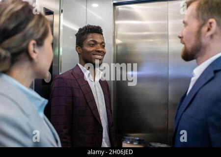 Happy young African businessman smiling while looking at male colleague during communication inside elevator Stock Photo