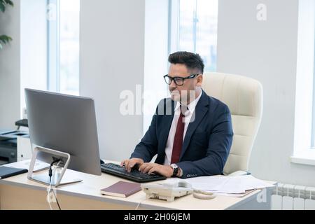 Busy director in elegant suit typing and looking at computer screen while sitting by desk in office Stock Photo