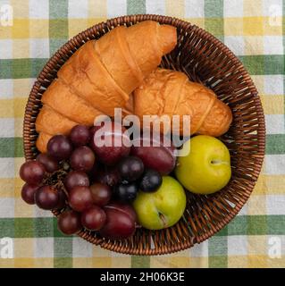 top view of fruits as grape pluots sloe berries with croissants in basket on plaid cloth background Stock Photo