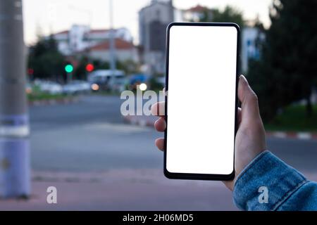 Woman using black smartphone. Mockup image of woman hand holding mobile phone with blank white screen, blurry city view in the background. Stock Photo