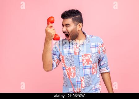 Portrait of bearded angry man screaming into phone handset, annoyed by telephone conversation, aggressive customer calling contact center. Indoor studio shot isolated on pink background. Stock Photo