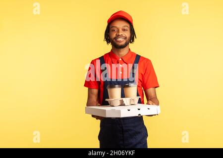 Delivery service. Friendly professional courier in blue overalls holding coffee and pizza box, wearing, offering drinks and food, looking at camera. Indoor studio shot isolated on yellow background. Stock Photo