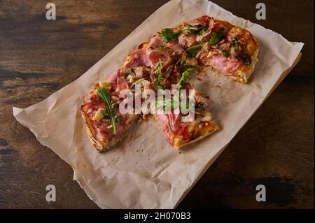 Cut into pieces rectangular Roman pizza with prosciutto ham, tomatoes, mozzarella and arugula on on paper on wooden background Stock Photo