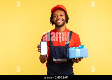 Portrait of happy smiling deliveryman in blue overalls, red T-shirt and cap holding present box and smart phone with blank screen for advertisement. Indoor studio shot isolated on yellow background. Stock Photo