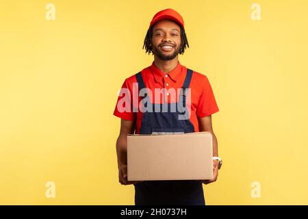 Smiling positive courier wearing blue overalls holding cardboard parcel, delivering orders to address, looking at camera with friendly expression. Indoor studio shot isolated on yellow background. Stock Photo
