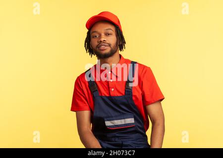 Portrait of bearded handyman with pleasant appearance wearing blue overalls, res T-shirt and visor cap, posing looking directly at camera. Indoor studio shot isolated on yellow background. Stock Photo