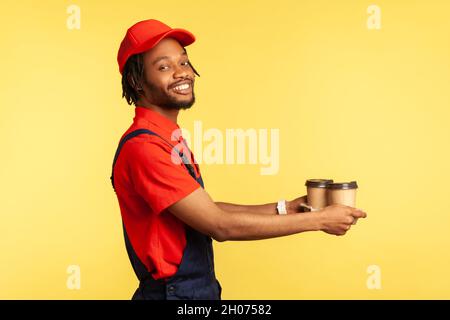 Side view portrait of smiling positive courier man wearing red T-shirt and blue uniform, giving take away coffee to client, delivery order. Indoor studio shot isolated on yellow background. Stock Photo