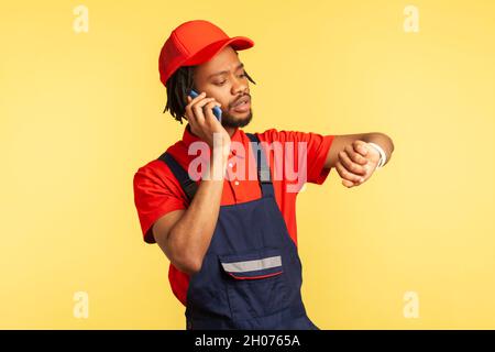 Portrait of concentrated courier wearing blue overalls talking phone with client, looking at smart watch, delivery service on time. Indoor studio shot isolated on yellow background. Stock Photo