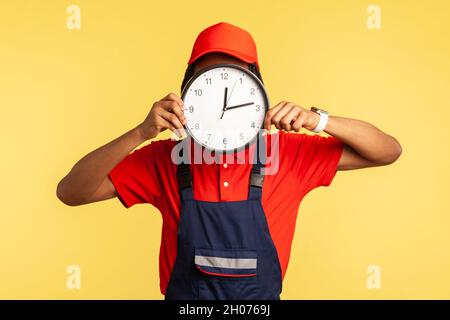 Anonymous labourer in uniform covering face with clock, advertising professional house repair services on time, punctual courier delivery. Indoor studio shot isolated on yellow background. Stock Photo