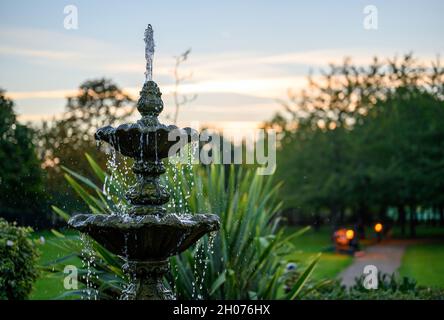 Fountain with water drops in a park with trees behind at dusk. The fountain is seen with a path, bench and lights just after sunset. Stock Photo