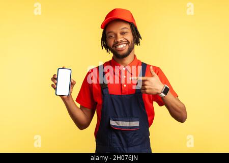 Portrait of happy handyman wearing uniform pointing at smart phone with blank screen, advertising new service industry application. Indoor studio shot isolated on yellow background. Stock Photo