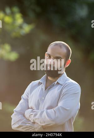 Handsome young man with beard standing and holding crossed arms in park at sunset Stock Photo