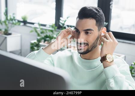 Smiling arabian businessman holding wireless earphone near blurred computer monitor in office Stock Photo