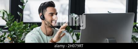 Smiling muslim businessman in headset having video call on computer in office, banner Stock Photo
