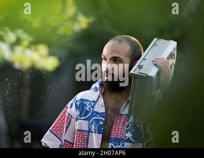 Young handsome man with beard holding vintage tape recorder on shoulder and walking in park at sunset Stock Photo
