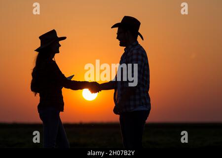 Silhouettes of man and woman farmers with hats shaking hands in field during sunset Stock Photo