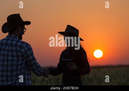Silhouettes of man and woman farmers with hats shaking hands in field during sunset Stock Photo