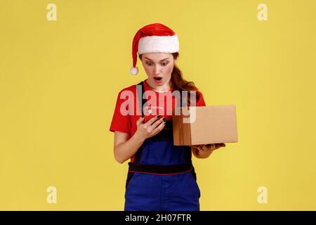 Surprised delivery woman with parcel in hands, using cell phone, having mistake with client address, wearing blue overalls and santa claus hat. Indoor studio shot isolated on yellow background. Stock Photo