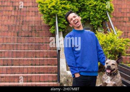 Young Latino man wearing blue clothes laughing loudly next to his pet pitbull. Harmless brindle colored dog. Happy man with his dog. Stock Photo