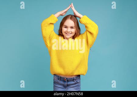 Little girl doing house roof gesture with hands over head and laughing, child care and protection, wearing yellow casual style sweater. Indoor studio shot isolated on blue background. Stock Photo