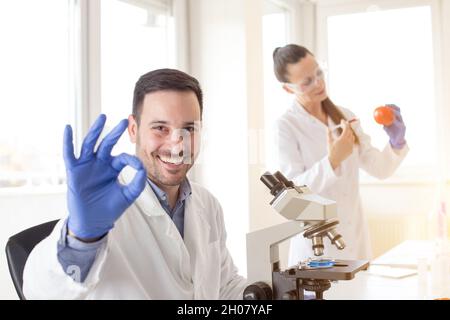 Satisfied scientist at microscope showing ok sign while woman biologist working in background Stock Photo