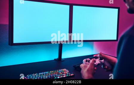 Over shoulder view of man holding game controller and playing on dual display. Blank monitor to insert your content. Professional computer game playin Stock Photo