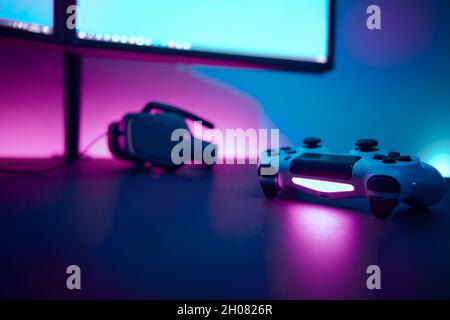 Gaming controller laying on dark table in front of dual lcd display. Light blue and pink illuminated wall in background. Professional computer game pl Stock Photo