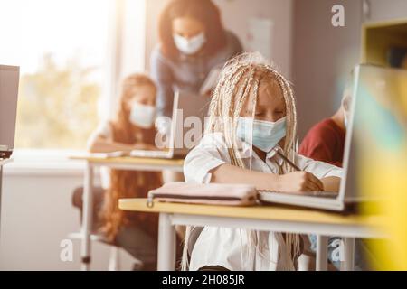 Schoolgirl in mask sitting at her desk and making notes Stock Photo
