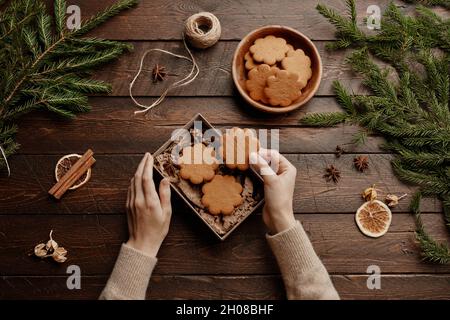 Top view Christmas background with unrecognizable young woman packing homemade cookies in gift wrap at wooden table, copy space Stock Photo
