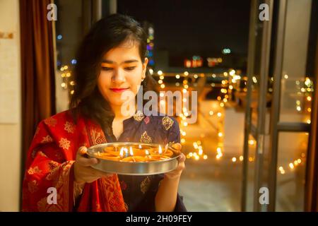 Happy Young indian smiling beautiful woman holding plate/thali with diya/clay oil lamps wearing traditional dress,standing indoor.Gorgeous girl in eth Stock Photo