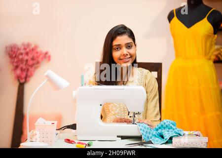 Portrait of Young indian happy woman,tailor,fashion designer,handmade dress maker using sewing machine,sitting behind her desk in her garment tailorin Stock Photo