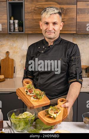 The chef is holding Italian bruschetta with cheese and figs. A stylish chef in a black jacket poses in his kitchen. Chef of Caucasian ethnicity prepar Stock Photo