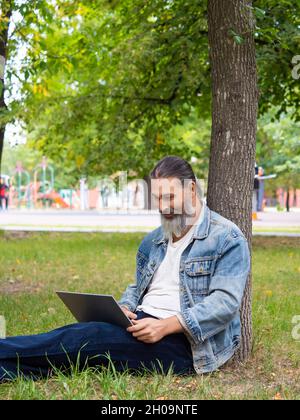 Photo of a bearded man while working online with a laptop. He sits in the park on the grass and works remotely. Vertrical photo. Stock Photo