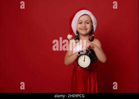 Beautiful adorable baby girl dressed in Santa carnival costume poses with alarm clock with midnight on face clock, looking at camera, against red colo Stock Photo
