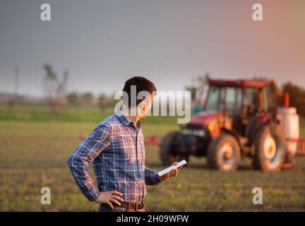 Young attractive farmer with tablet standing in front of tractor in field Stock Photo